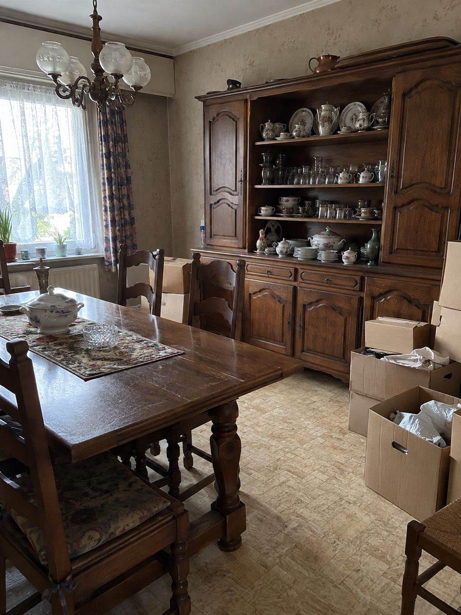 Salle à manger d’une maison des années 1960 avec une lourde table et des chaises en bois, un vaisselier rempli de vaisselle et des cartons empilés au sol
