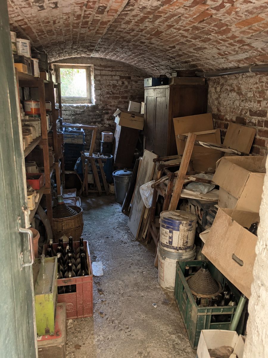 Cave voûtée en brique encombrée de vieilles étagères, de pots de peinture, de casiers de bouteilles, de meubles cassés et de cartons