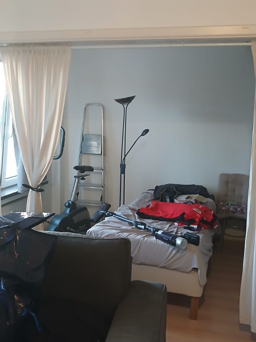 Chambre d’un appartement à Mons avec lit, vélo d’appartement, escabeau, lampadaire et canapé encore en place avant l’état des lieux