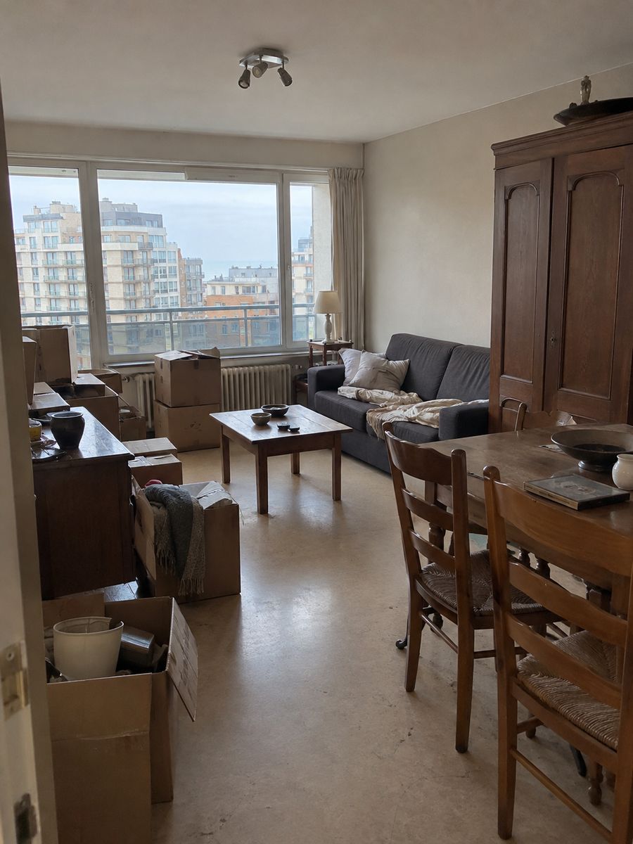 Séjour d’un appartement en étage à Ostende avec grandes fenêtres, canapé, garde-robe, table à manger et cartons prêts à être évacués